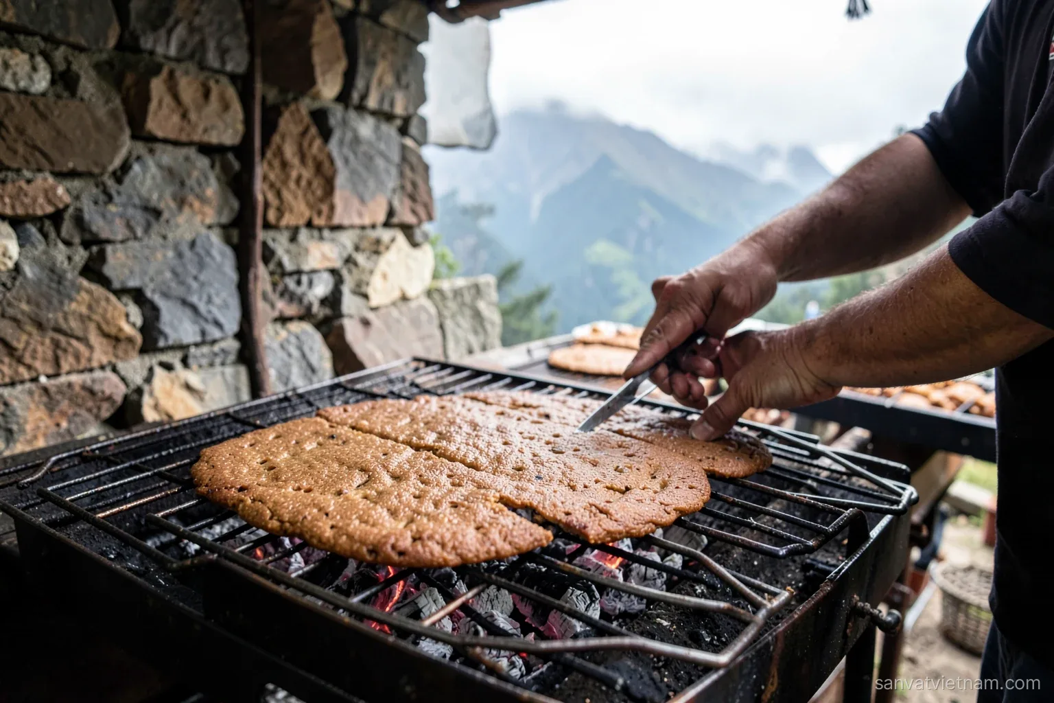 Round flat brown cakes toasting on a metal grill over charcoal at a highland market stall, a vendor's hands turning them, stone walls and misty peaks behind